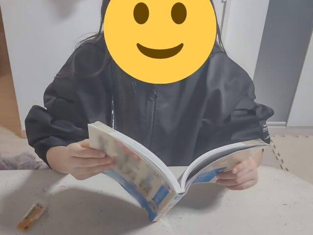 小学3年生の娘がカルパスを食べながらどんどん強くなるこども将棋勝てる手筋がわかる本を読んでいるところの写真